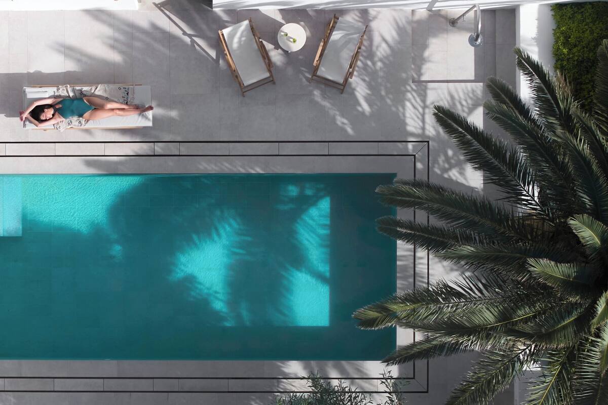An aerial view of a swimming pool with a person sunbathing, lounge chairs, and a small table.