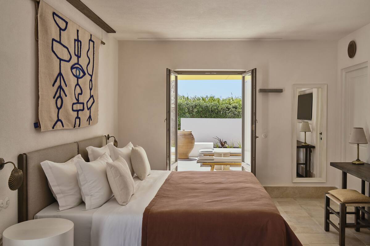 A bedroom with a large bed, a bedside table, a nightstand, a mirror, and a view of an outdoor patio with seating and a large vase.