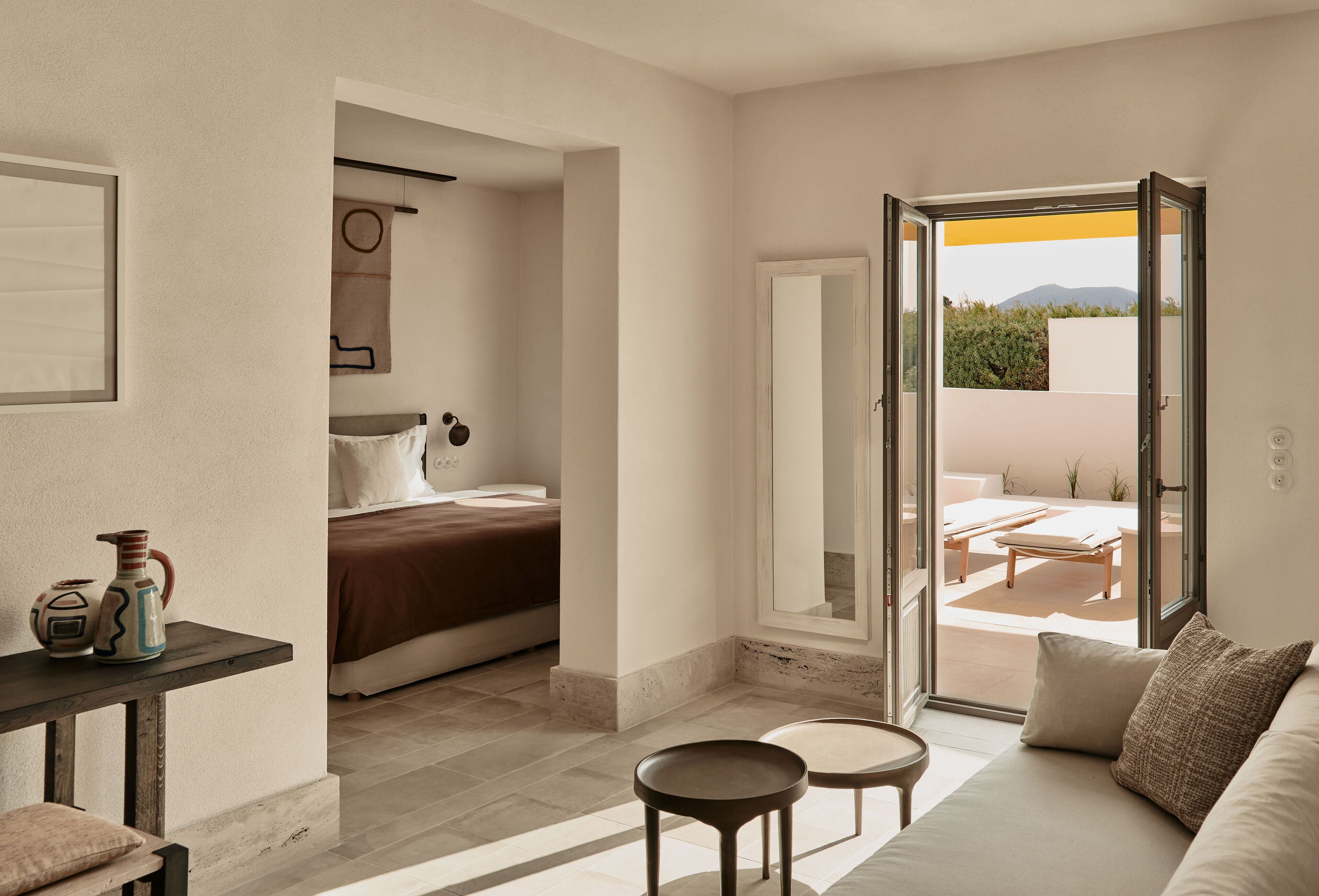 A modern hotel room with a bed, a small table, a sofa, and a balcony with outdoor furniture.
