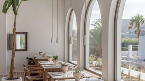 Un restaurant doté d’une longue table, de chaises, de plantes en pot et de grandes fenêtres offrant une vue sur des palmiers et un paysage lointain.