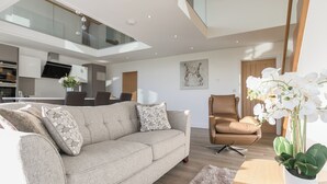 A modern living room with a sofa, a brown armchair, a dining area, and a kitchen in the background. - The Lookout - UKC3846 (Ticknall, near Derby)