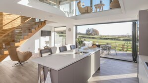 A modern kitchen with a large island, a dining area with chairs, a glass staircase, and a view of the countryside. - The Lookout - UKC3846 (Ticknall, near Derby)