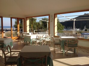 An outdoor dining area with tables and chairs, large windows with a view of the sea and boats, and a wooden pergola structure. - Hotel Villa Portuso (Monte Argentario)