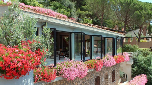 Ein Steinhaus mit einem Balkon, der mit rosa Blumen geschmückt ist, großen Fenstern und Blick auf die umliegende Grünfläche.