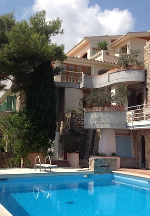 A poolside view of a multi-story building with balconies, a stone wall, and a large tree. - Hotel Villa Portuso (Monte Argentario)