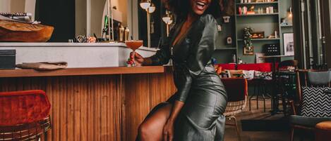 Une femme en robe, debout près d’un bar, dans un espace doté d’un sol en bois et d’un aménagement intérieur moderne.