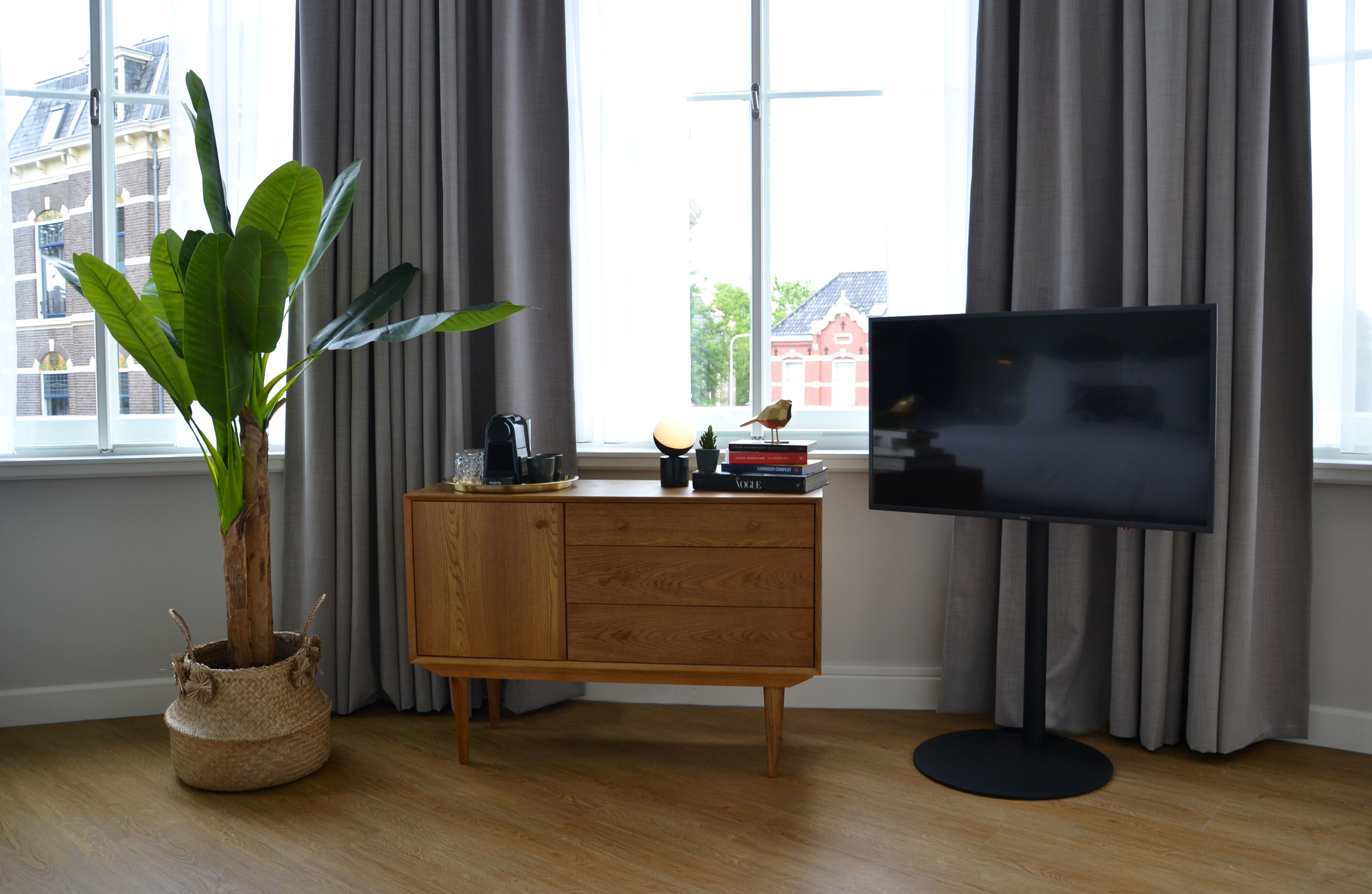 Una sala de estar moderna con un mueble de madera, un televisor, una planta en maceta y vistas de edificios a través de amplios ventanales.