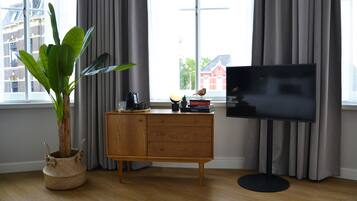 Una sala de estar moderna con un mueble de madera, un televisor, una planta en maceta y vistas de edificios a través de amplios ventanales.
