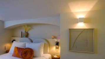 Een netjes opgemaakt bed met witte en oranje kussens, een nachtkastje met een lamp en een wandlamp.