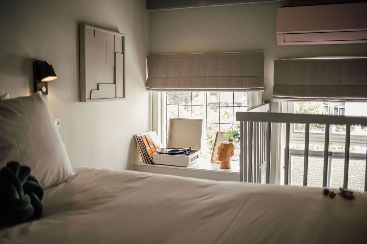 A bedroom with a bed, a bedside table with a record player, a lamp, and a window with a view of the outside.