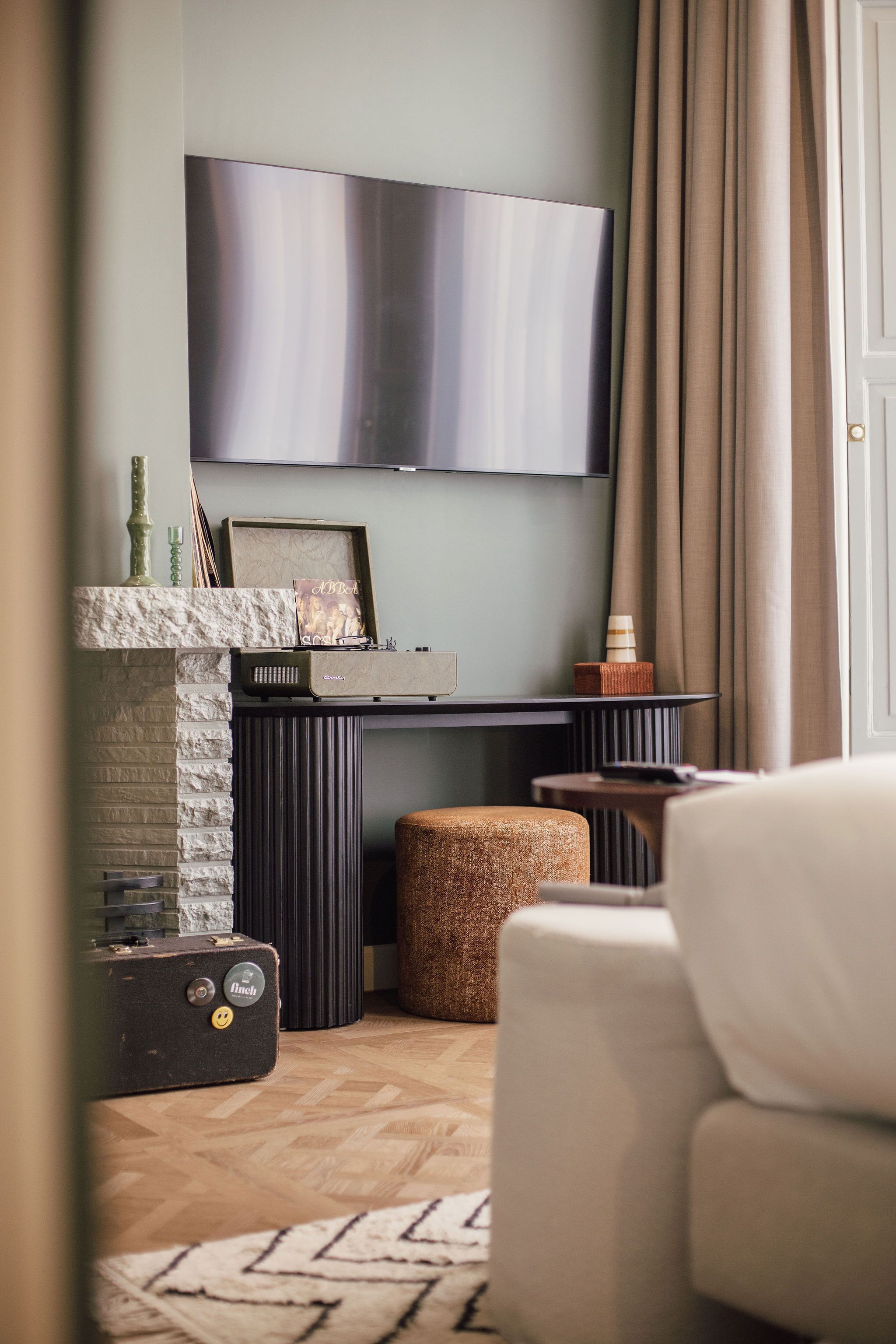 A living room with a flat-screen TV mounted on the wall, a fireplace with decorative items, a sofa, and a small table with a speaker.