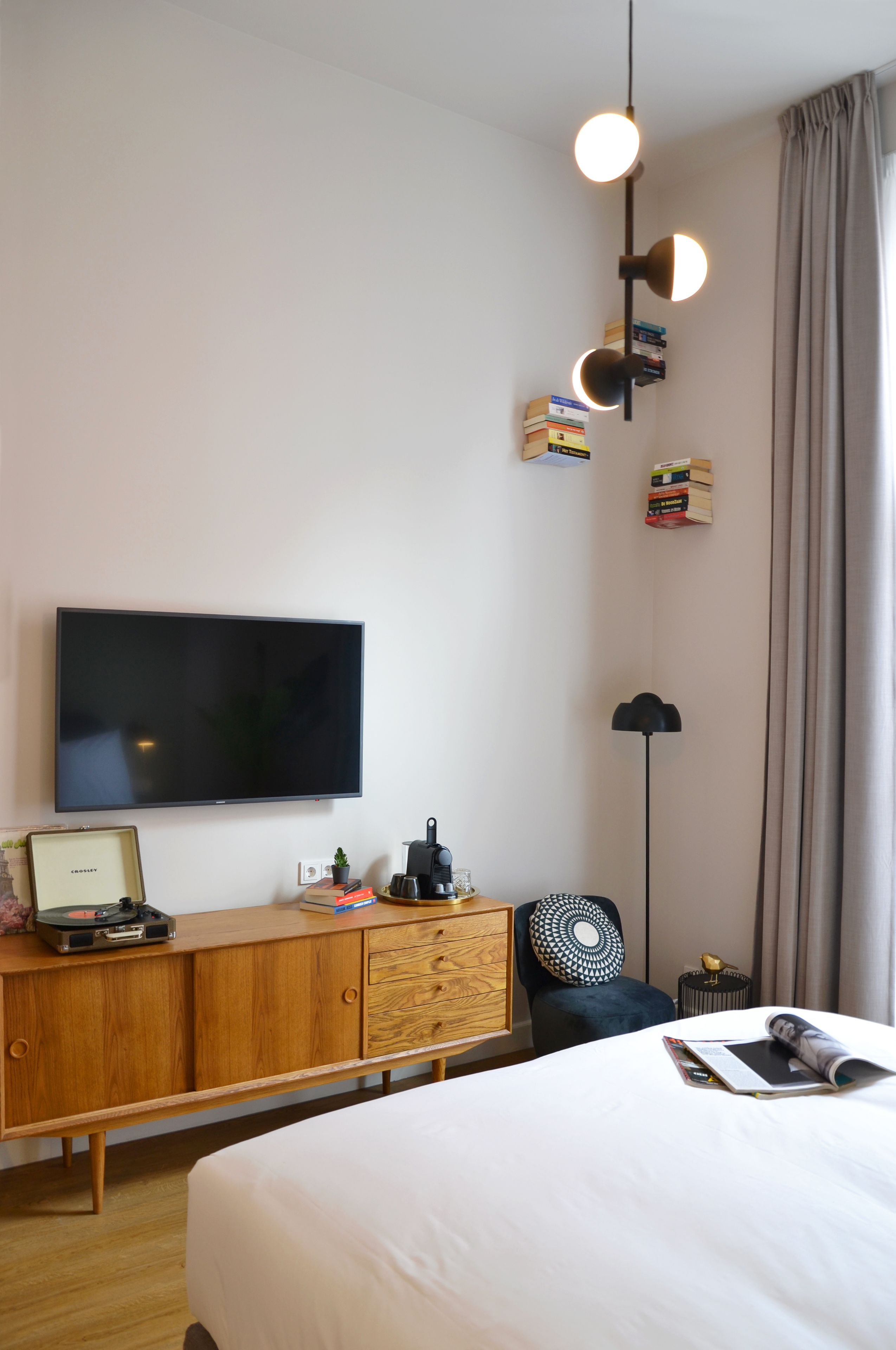 Una habitación de hotel moderna con televisión de pantalla plana, cómoda de madera, escritorio con lámpara, cama y un amplio ventanal con cortinas.
