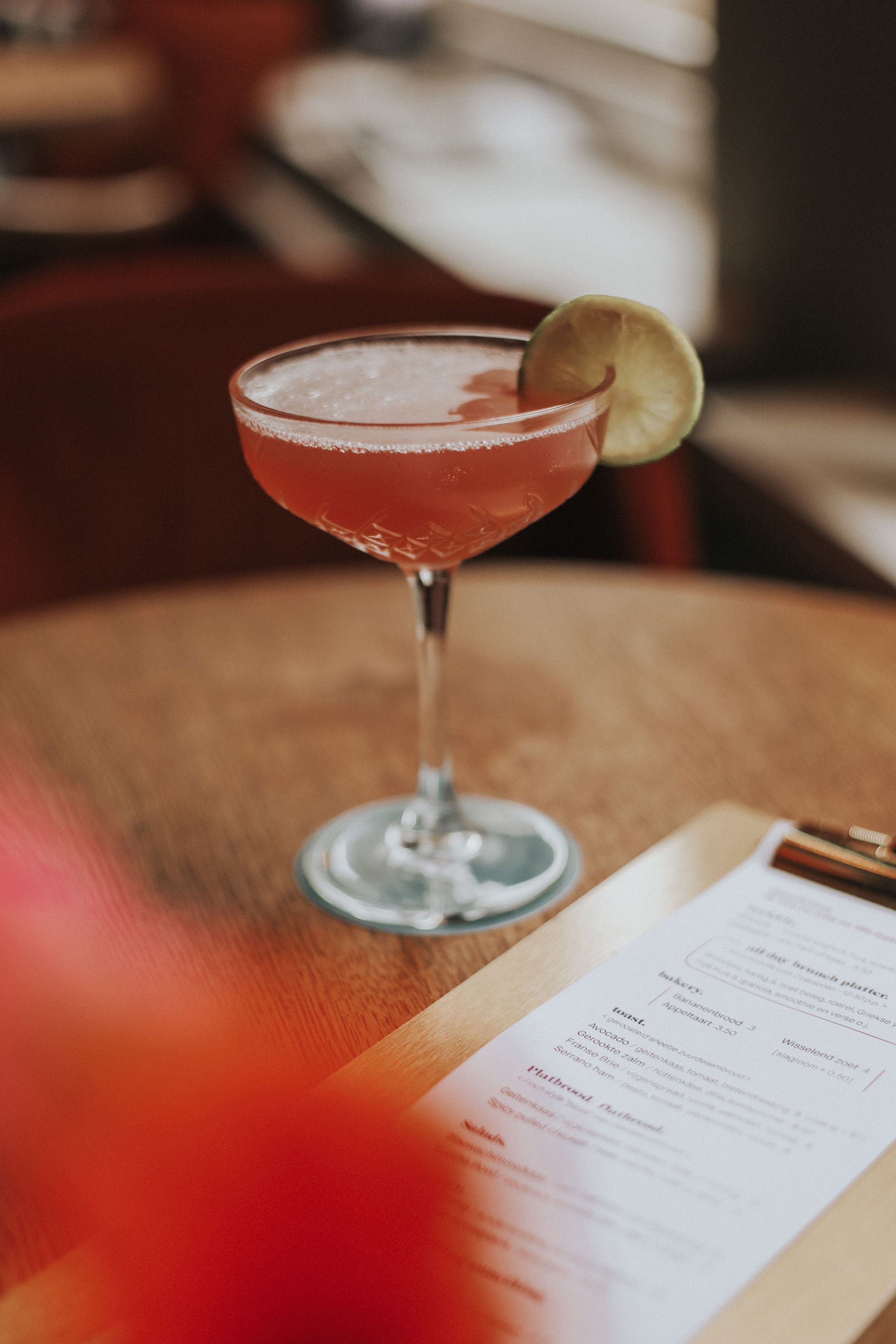 Un verre à cocktail, garni d’une tranche de citron vert, est posé sur une table en bois, et on aperçoit une partie d’un menu.