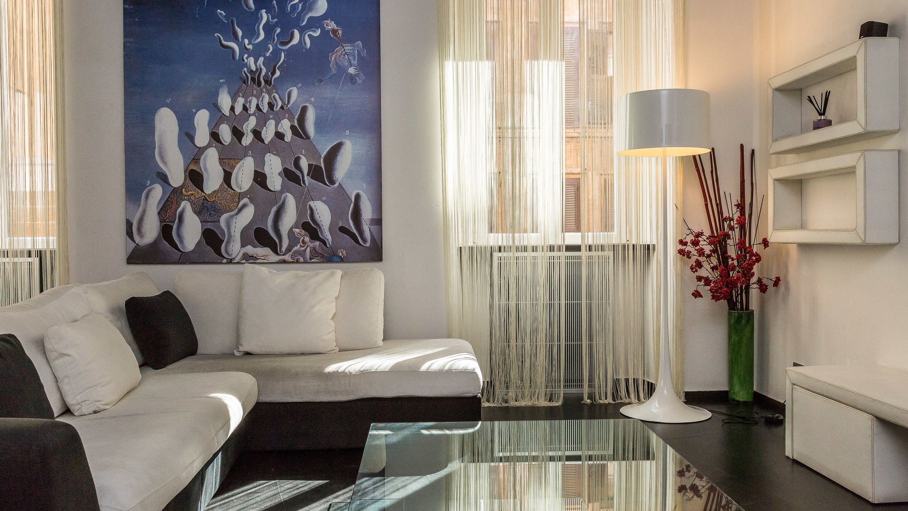 A modern living room with a sofa, a glass coffee table, a painting, a lamp, and a vase with red flowers.