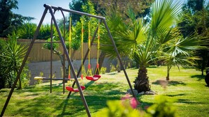 A swing set in a grassy backyard with palm trees and a wooden fence. - Denise Apartments (Zakynthos)