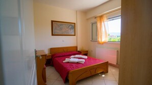 A bedroom with a wooden bed, a nightstand, a bedside table, a window with curtains, and a painting on the wall. - Denise Apartments (Zakynthos)