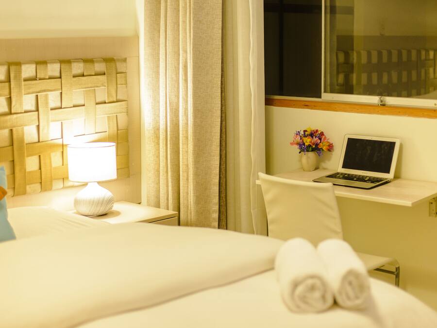 Quarto de hotel com cama, mesa de cabeceira com abajur, um laptop sobre uma pequena mesa e uma janela com cortinas.