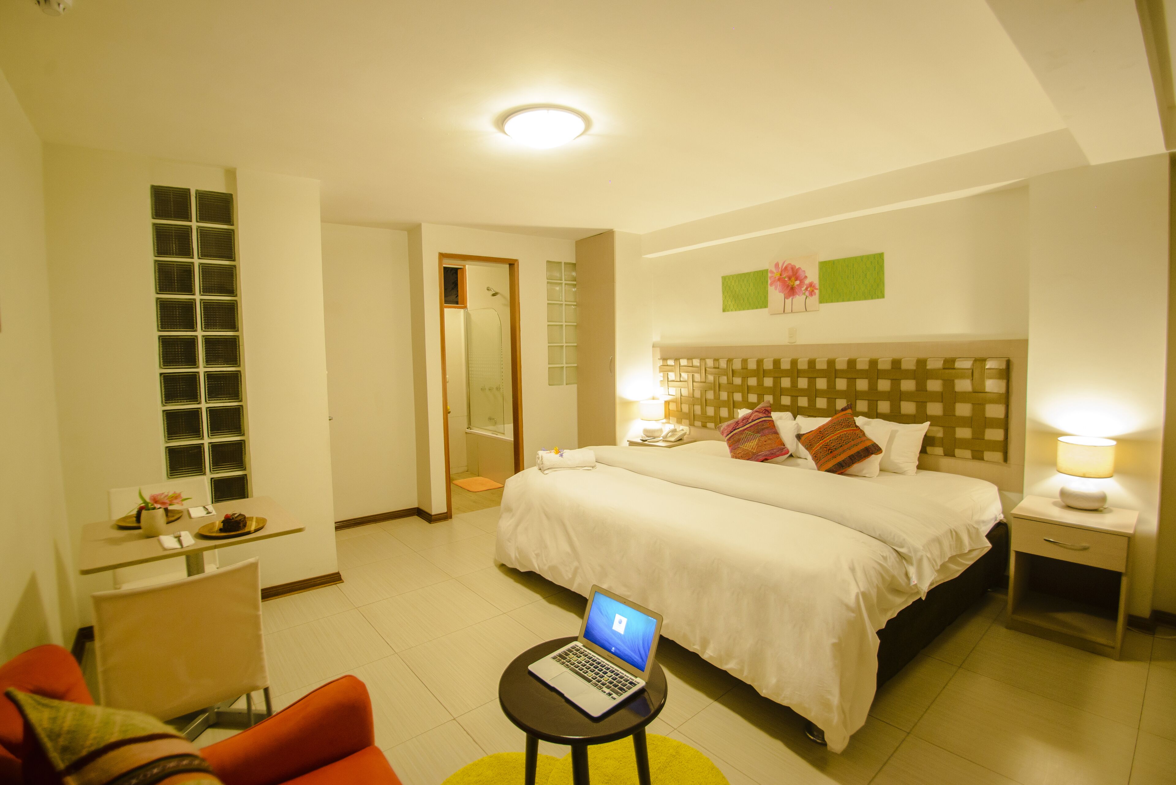 Habitació d'hotel amb un llit, escriptori amb un ordinador portàtil, cadira, taulella de nit amb una làmpada i una taula petita amb un ram i flors.