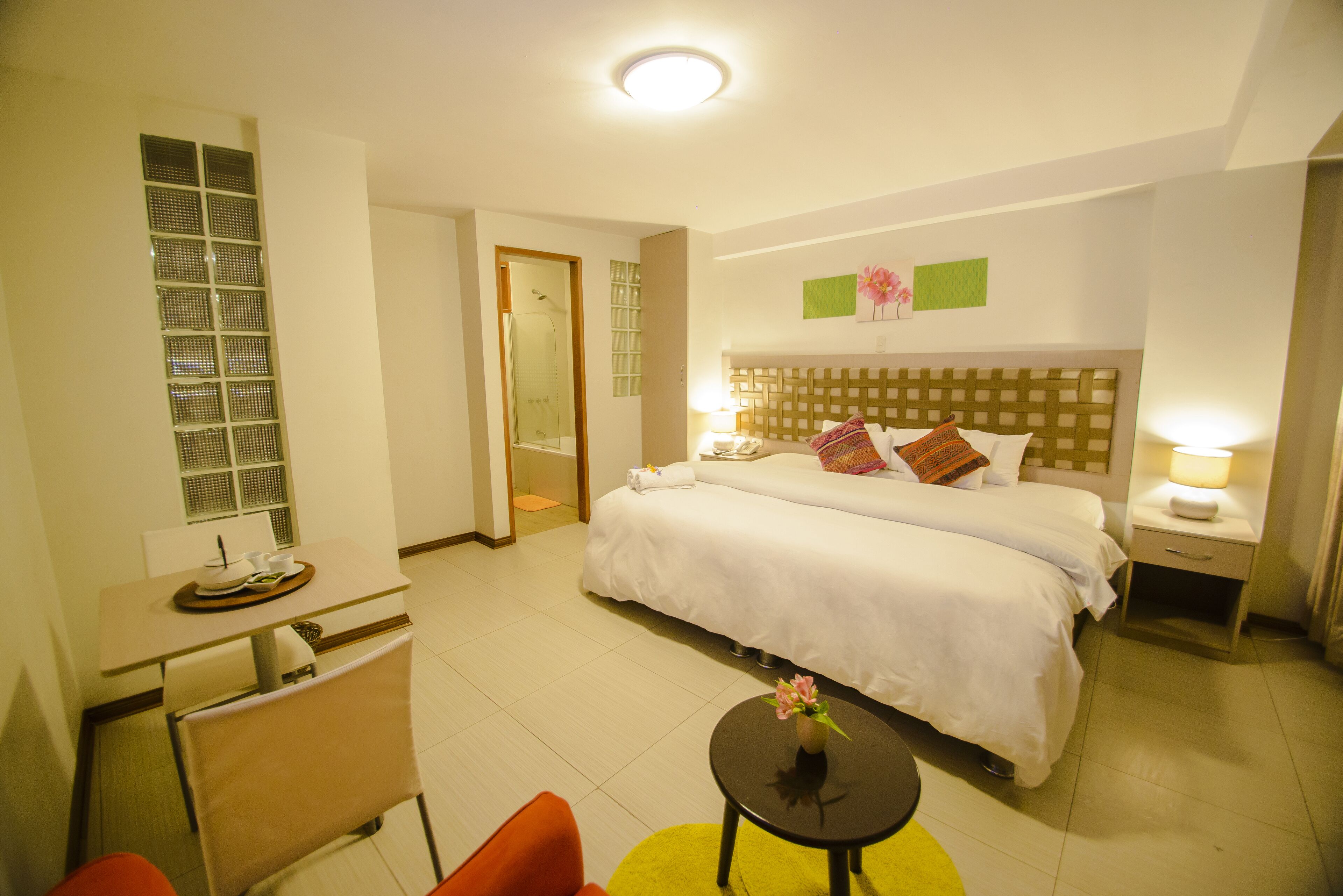A hotel room with a large bed, a desk with a chair, a small table with a vase, and a bathroom visible through an open door.
