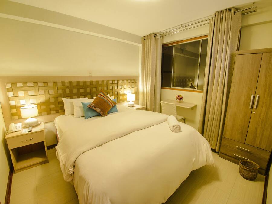 Habitación de hotel con una cama grande, una mesita de noche con lámpara, un armario y una ventana con cortinas.