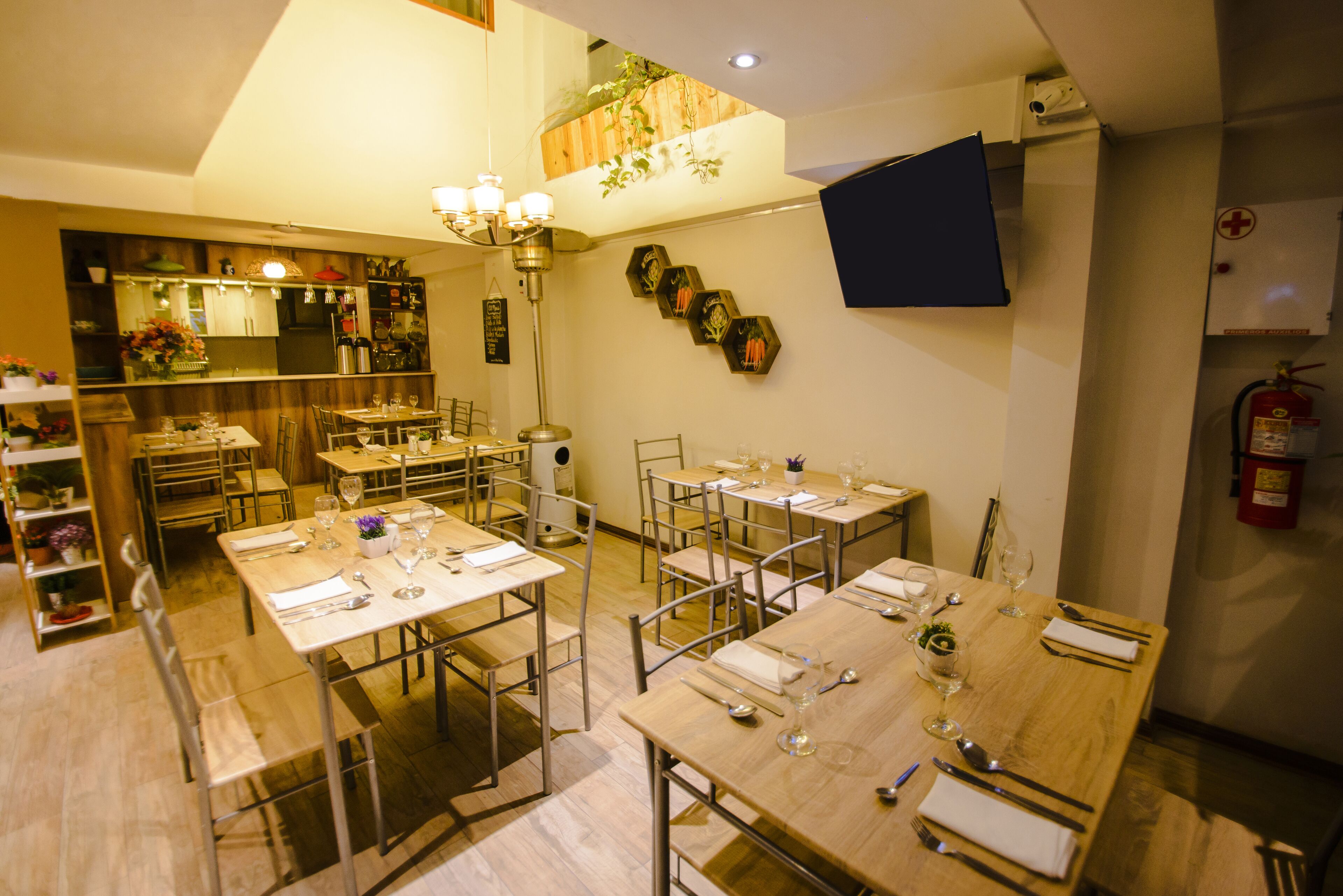 A dining area with multiple tables set for meals, a kitchen in the background, and a mounted TV on the wall.