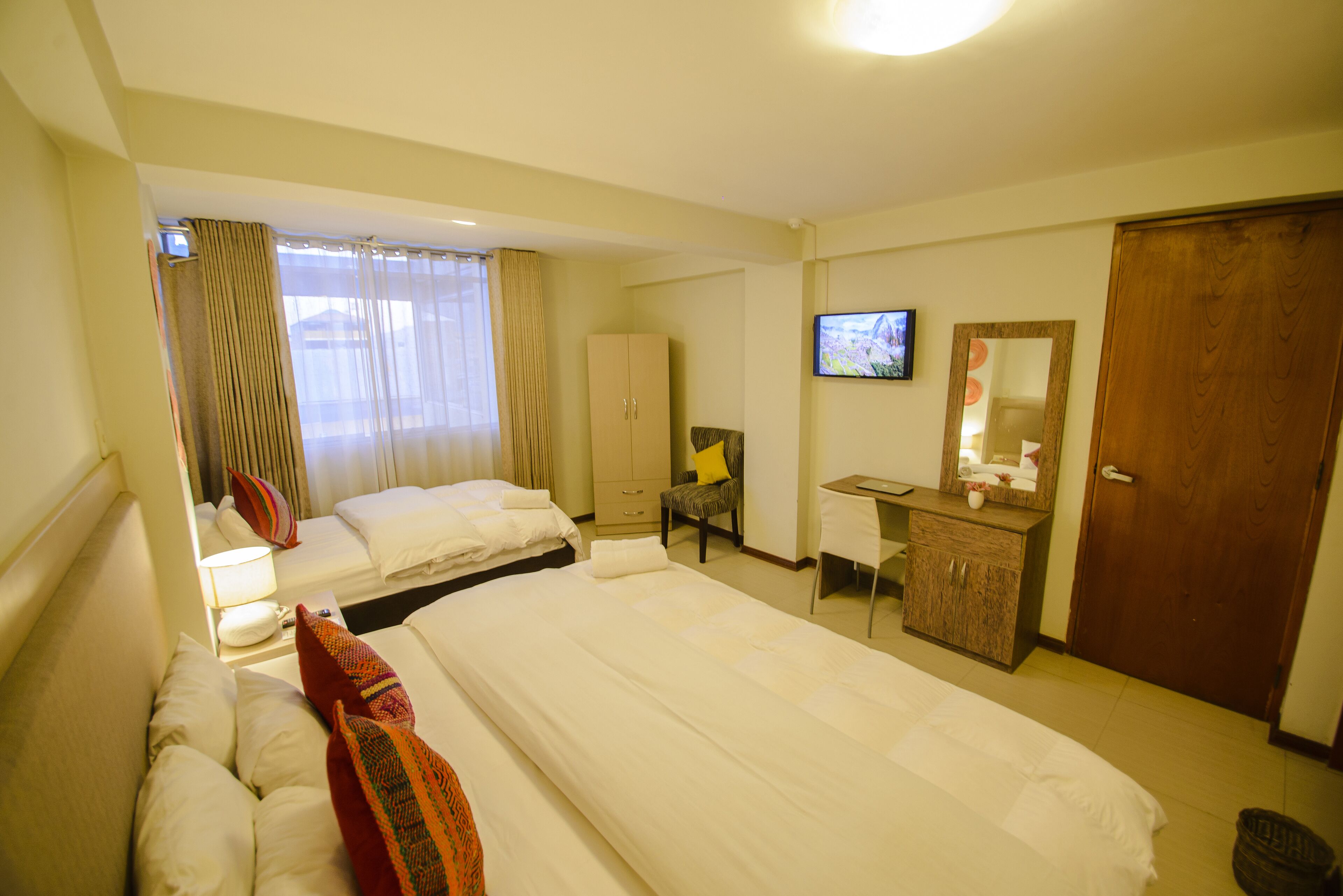 Quarto de hotel com duas camas, uma escrivaninha, uma cadeira, uma televisão e um guarda-roupa.