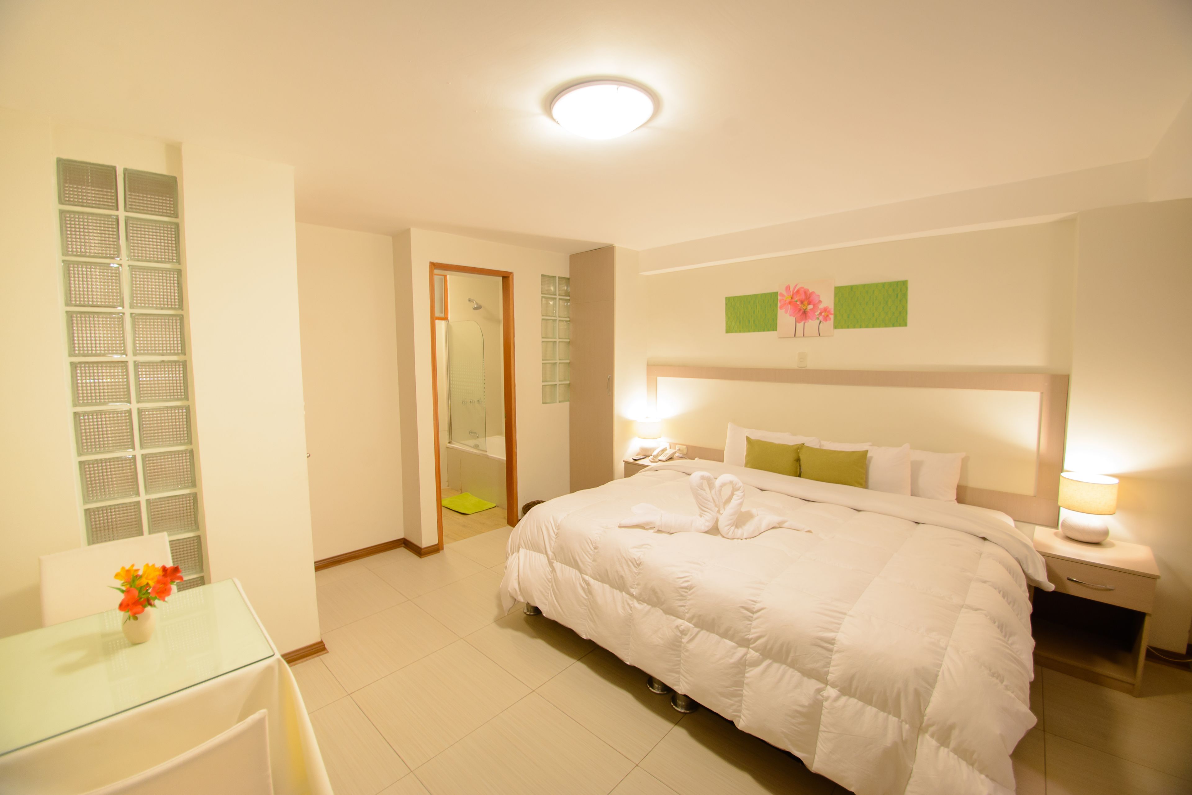 Una habitación de hotel con una cama grande, mesitas de noche, una mesa de vidrio con flores y una zona de ducha.