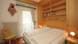 Quarto com cama, uma cadeira, prateleira com objetos decorativos e janela com cortinas.
