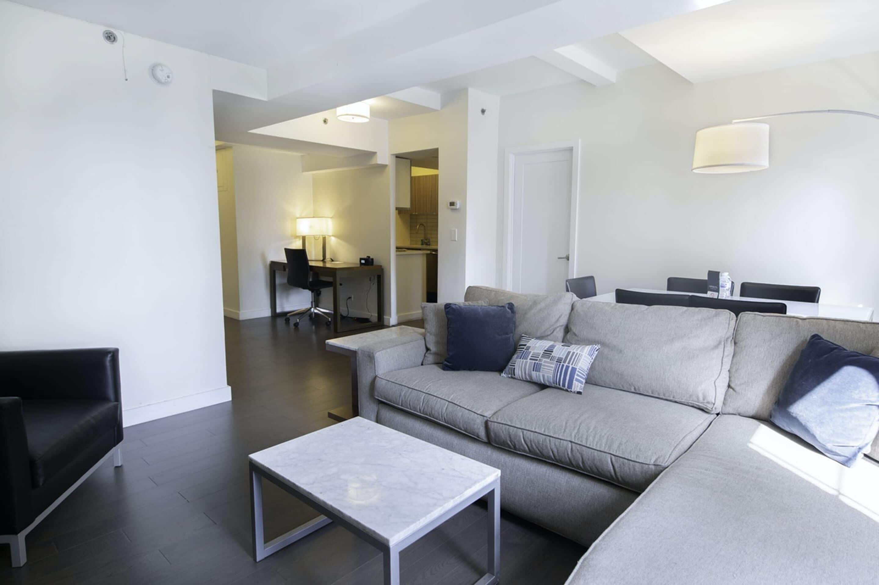 A modern living room with a grey sofa, a black armchair, and a small table. There is a kitchen in the background with a dining table and chairs.