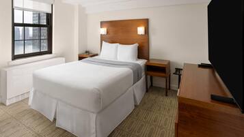 A hotel room with a large bed, a wooden headboard, two bedside tables, a TV, and a window with a view of buildings.