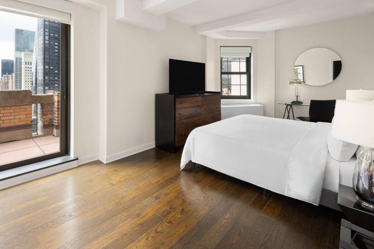 A modern hotel room with a large bed, a flat-screen TV, a dresser, and a desk with a lamp.