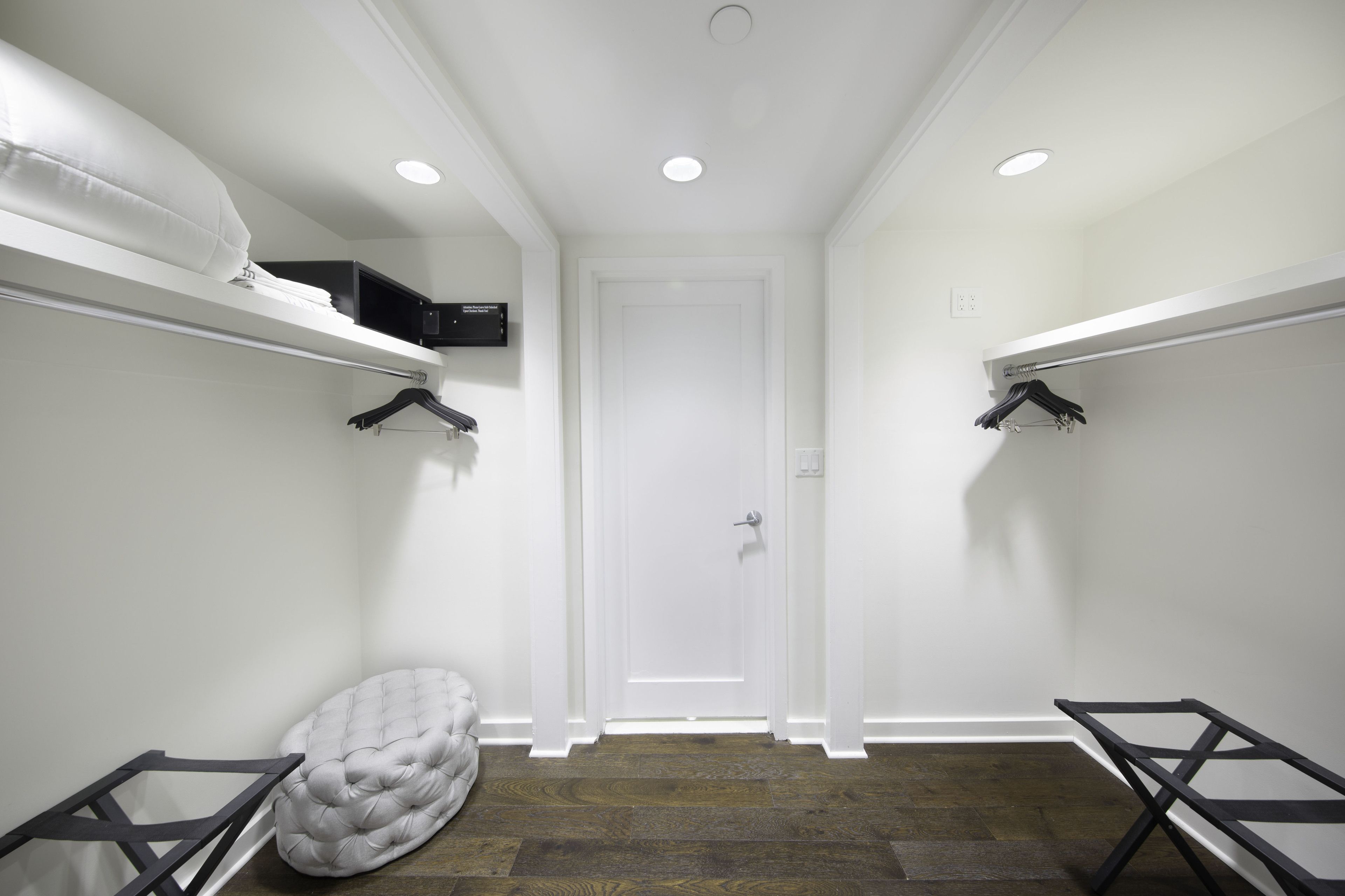 A narrow hallway with white walls, a white door at the end, and a small seating area with a tufted ottoman.