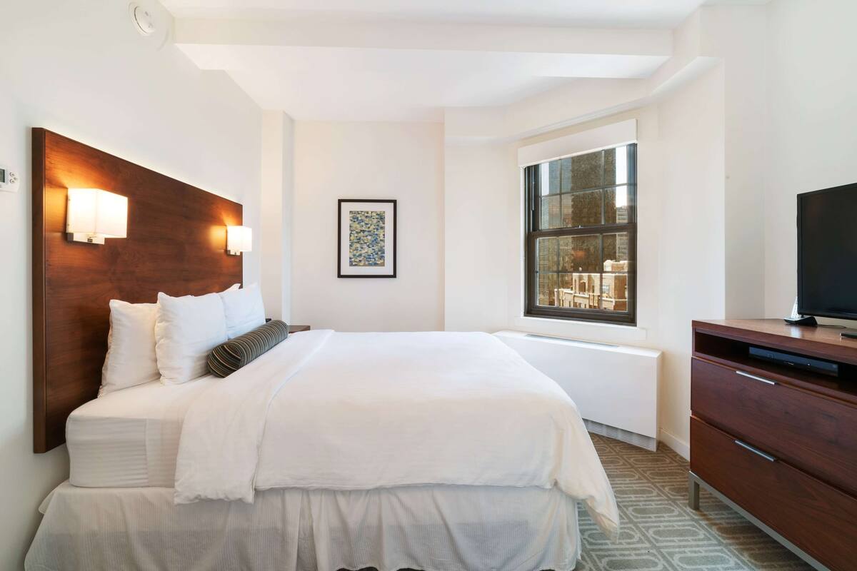 A modern hotel room with a large bed, bedside lamps, a window with a view, and a TV on a wooden cabinet.