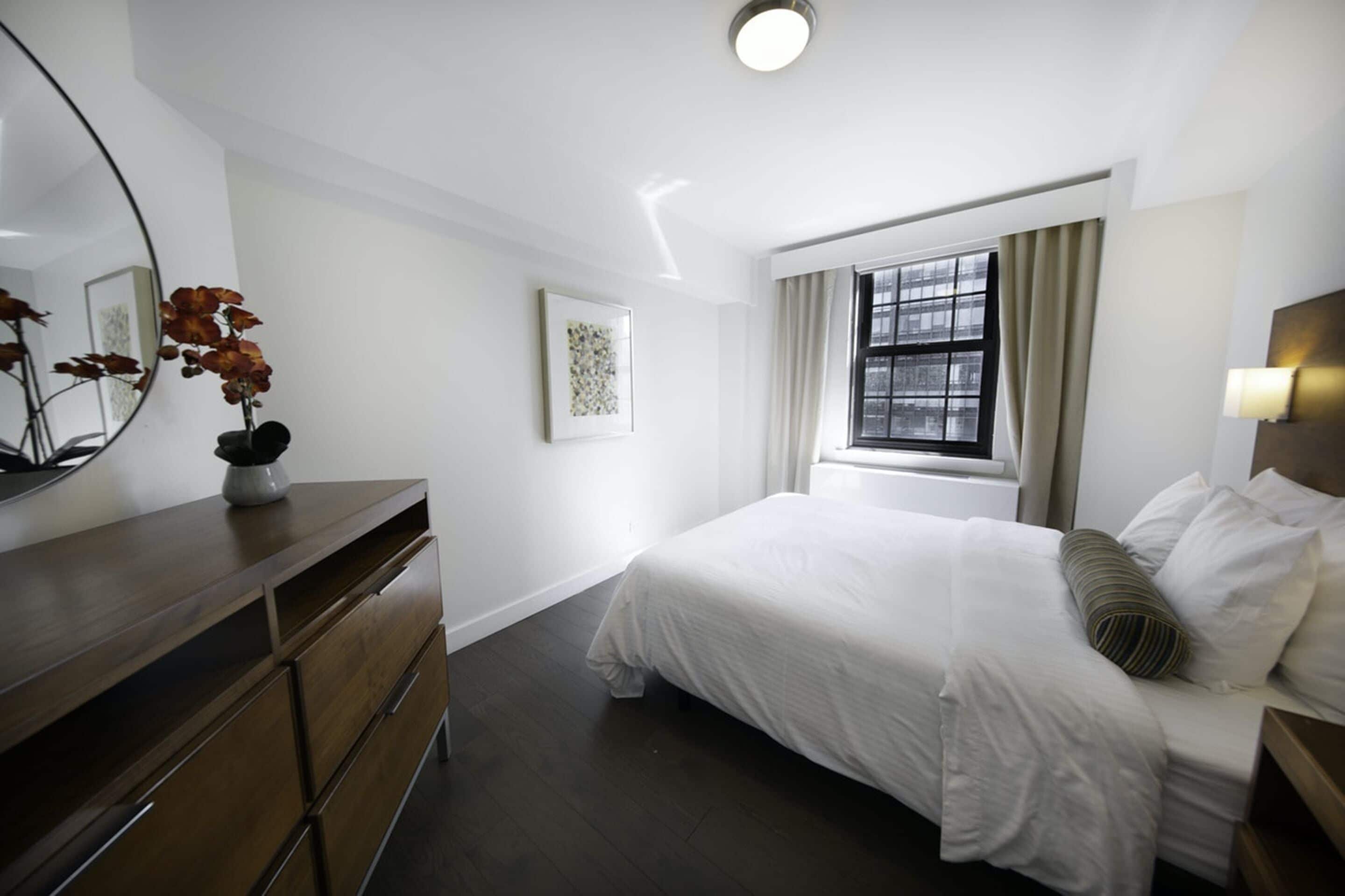 A hotel room with a bed, a dresser, a mirror, a window with curtains, and a wall-mounted light.