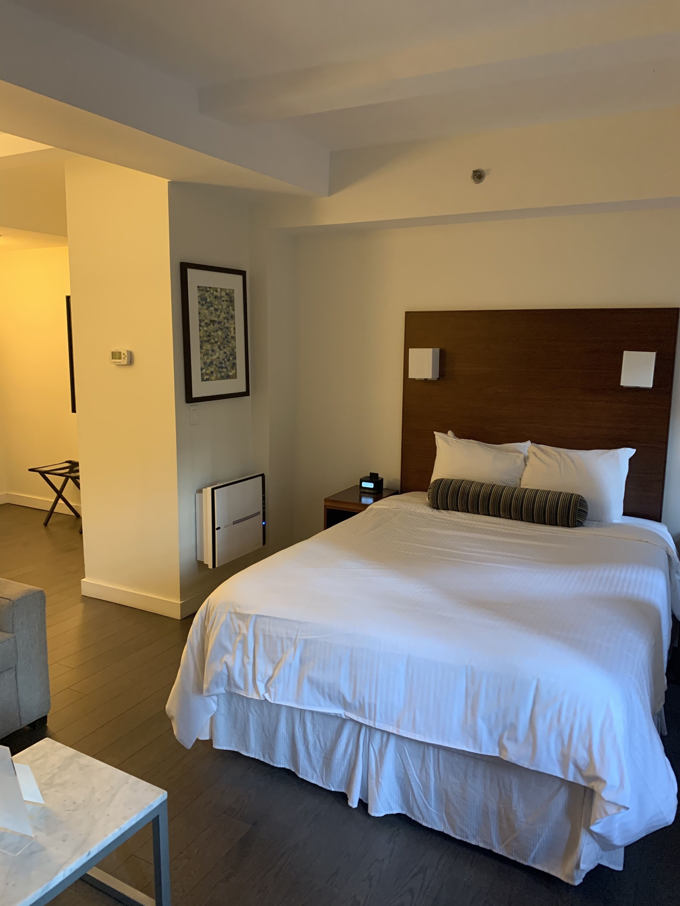 A hotel room with a large bed, a bedside table, a nightstand, a framed picture on the wall, and a small table with a chair.