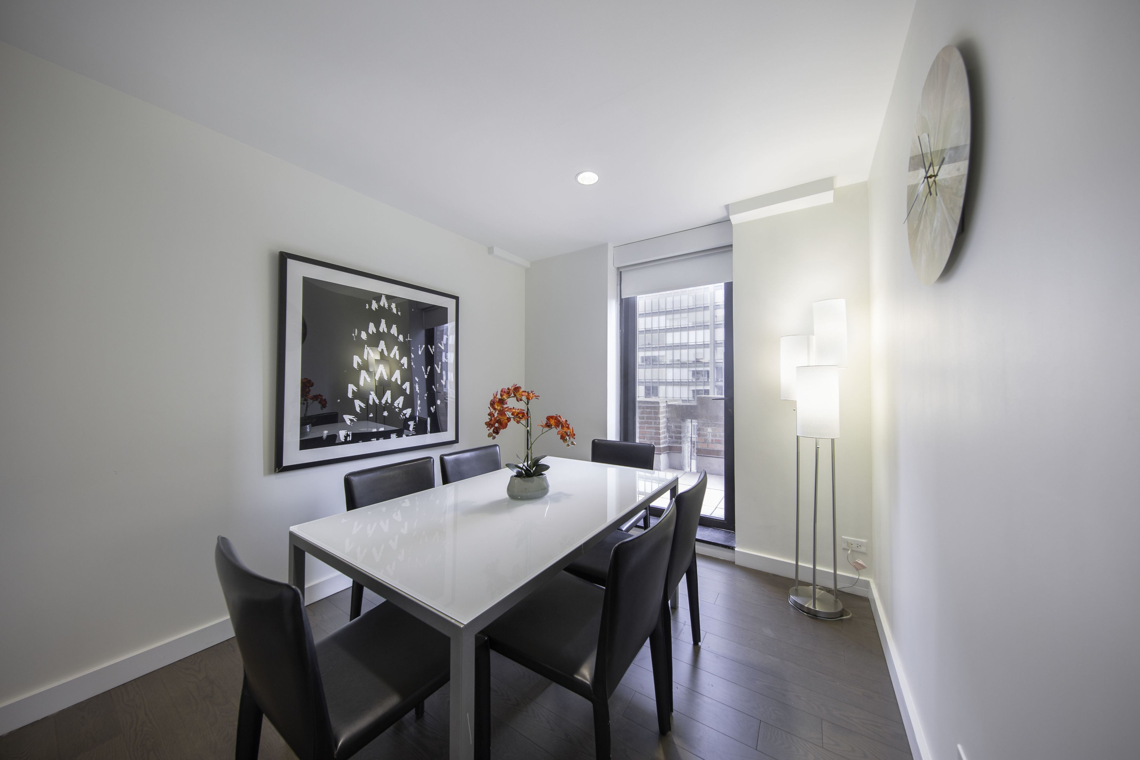 A modern dining area with a white table, black chairs, a vase with flowers, a framed abstract painting, and a floor lamp.