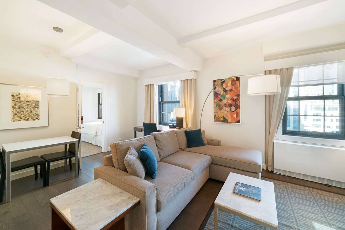 A modern hotel room with a sectional sofa, a small table, a desk, and a large window with curtains.