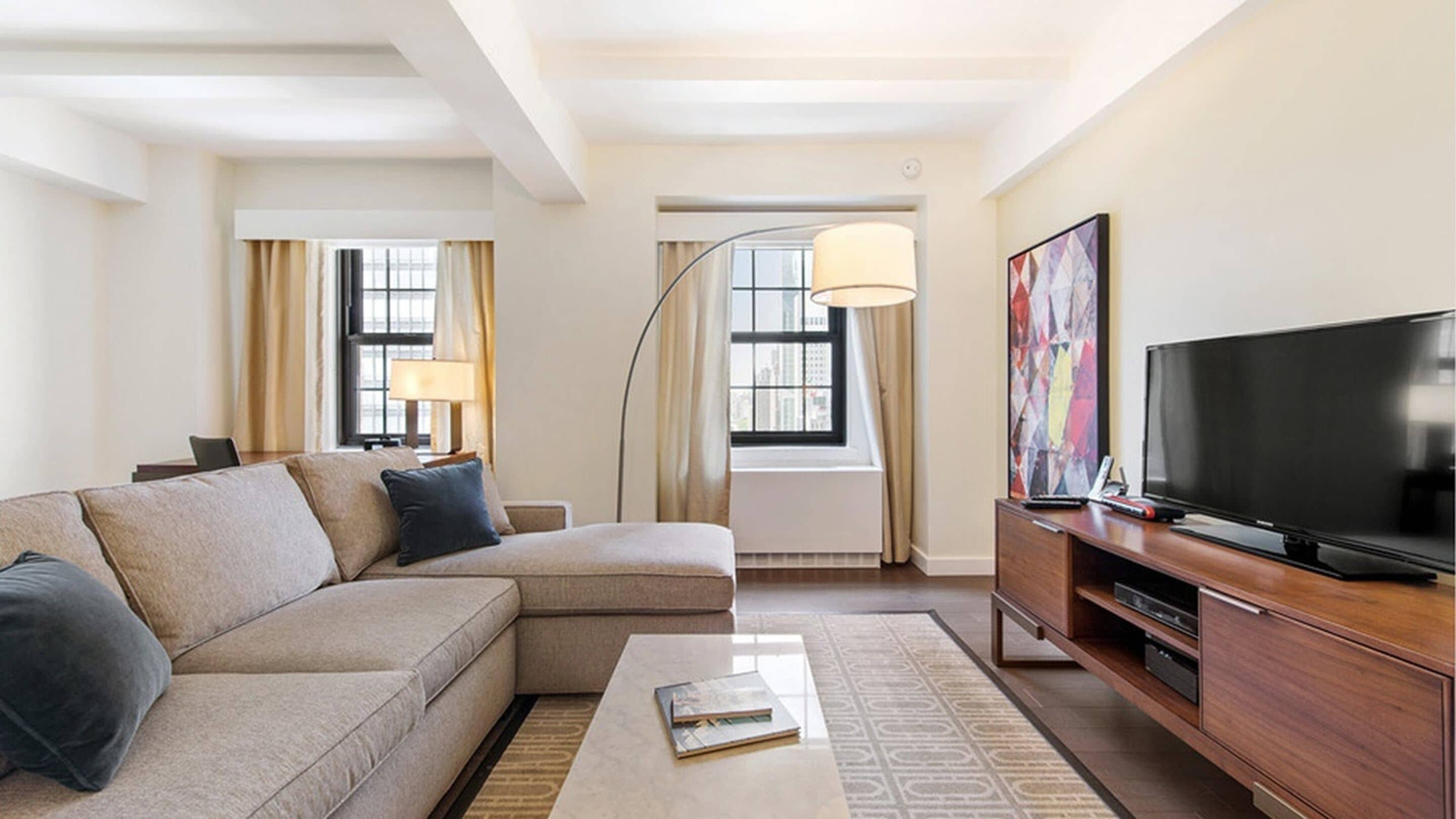 A modern hotel room with a sectional sofa, a flat-screen TV, a desk with a lamp, and a large window with curtains.