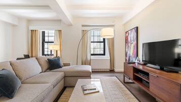 A modern hotel room with a sectional sofa, a flat-screen TV, a desk with a lamp, and a large window with curtains.