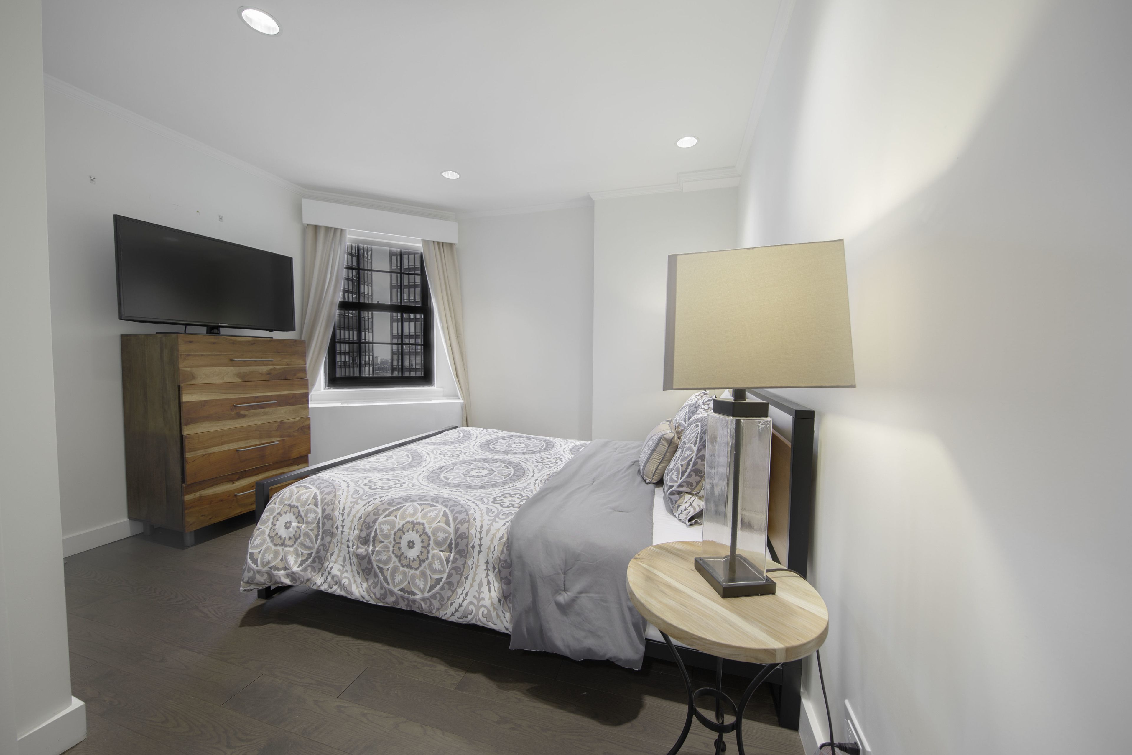 A bedroom with a bed, a TV, a dresser, a lamp, and a window with curtains.