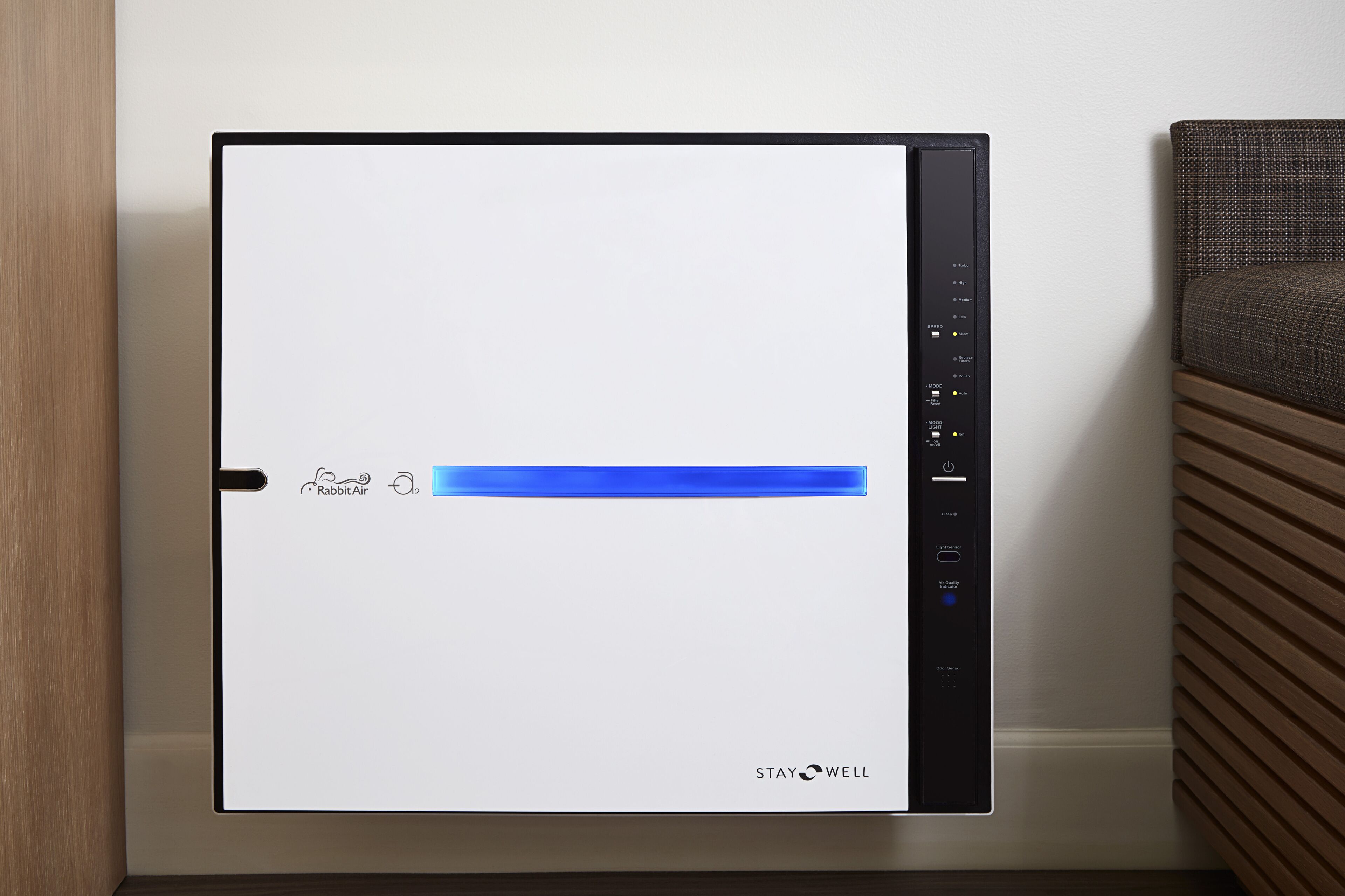 Un purificateur d’air moderne doté d’un panneau de commande, posé sur un plancher en bois, devant un mur blanc.
