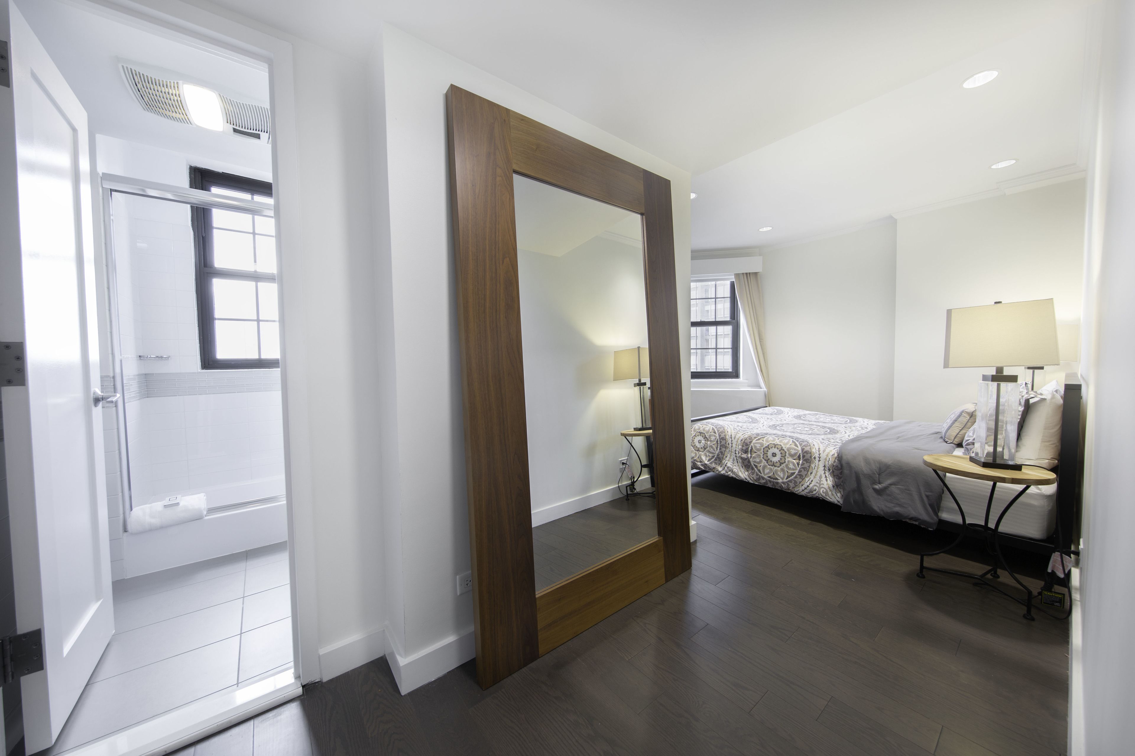 A hotel room with a large mirror, a bed with a patterned bedspread, a bedside table with a lamp, and a window with curtains.