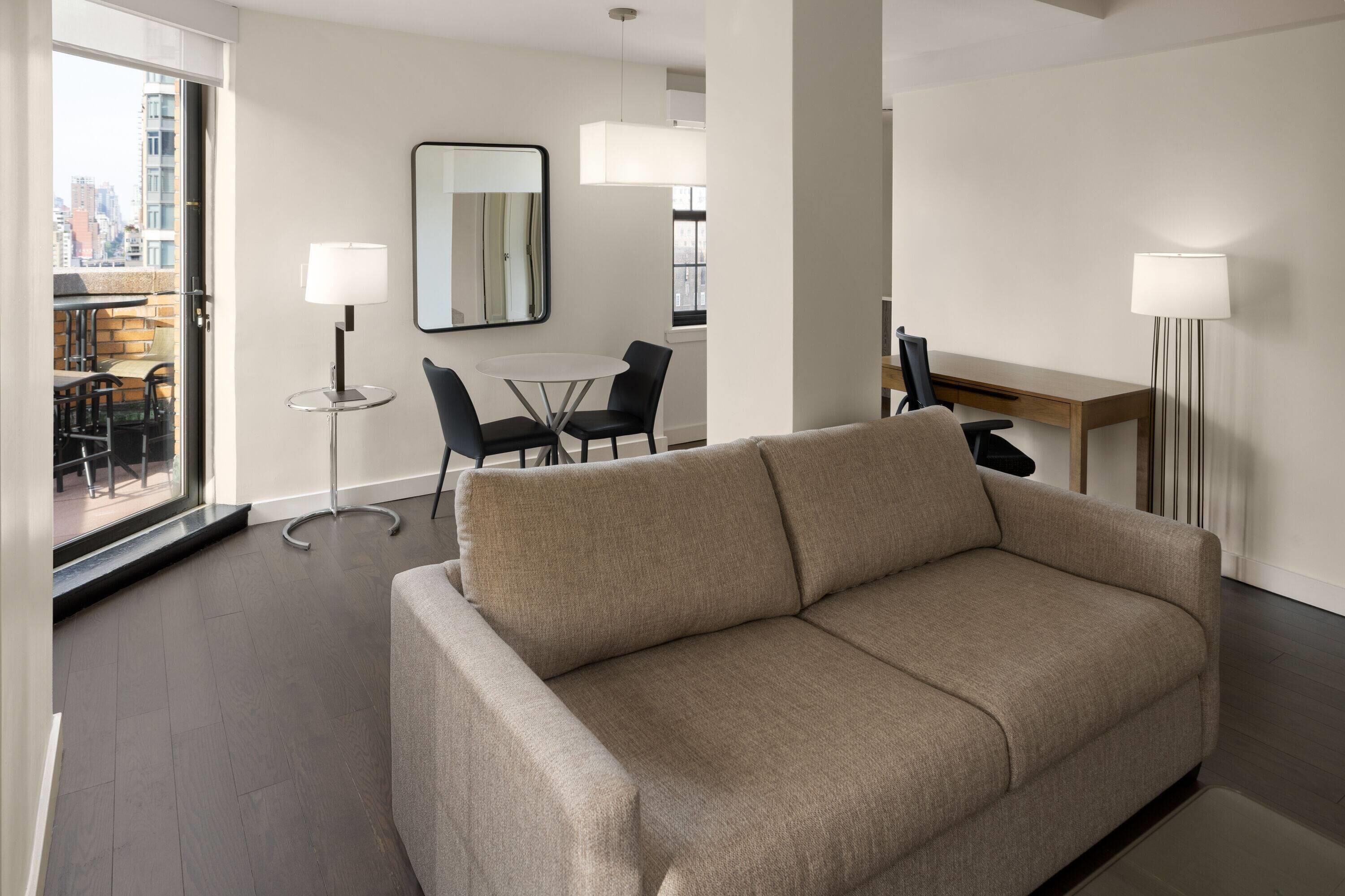 A modern hotel room with a beige sofa, a dining table with chairs, a small round table with a lamp, and a large mirror on the wall.