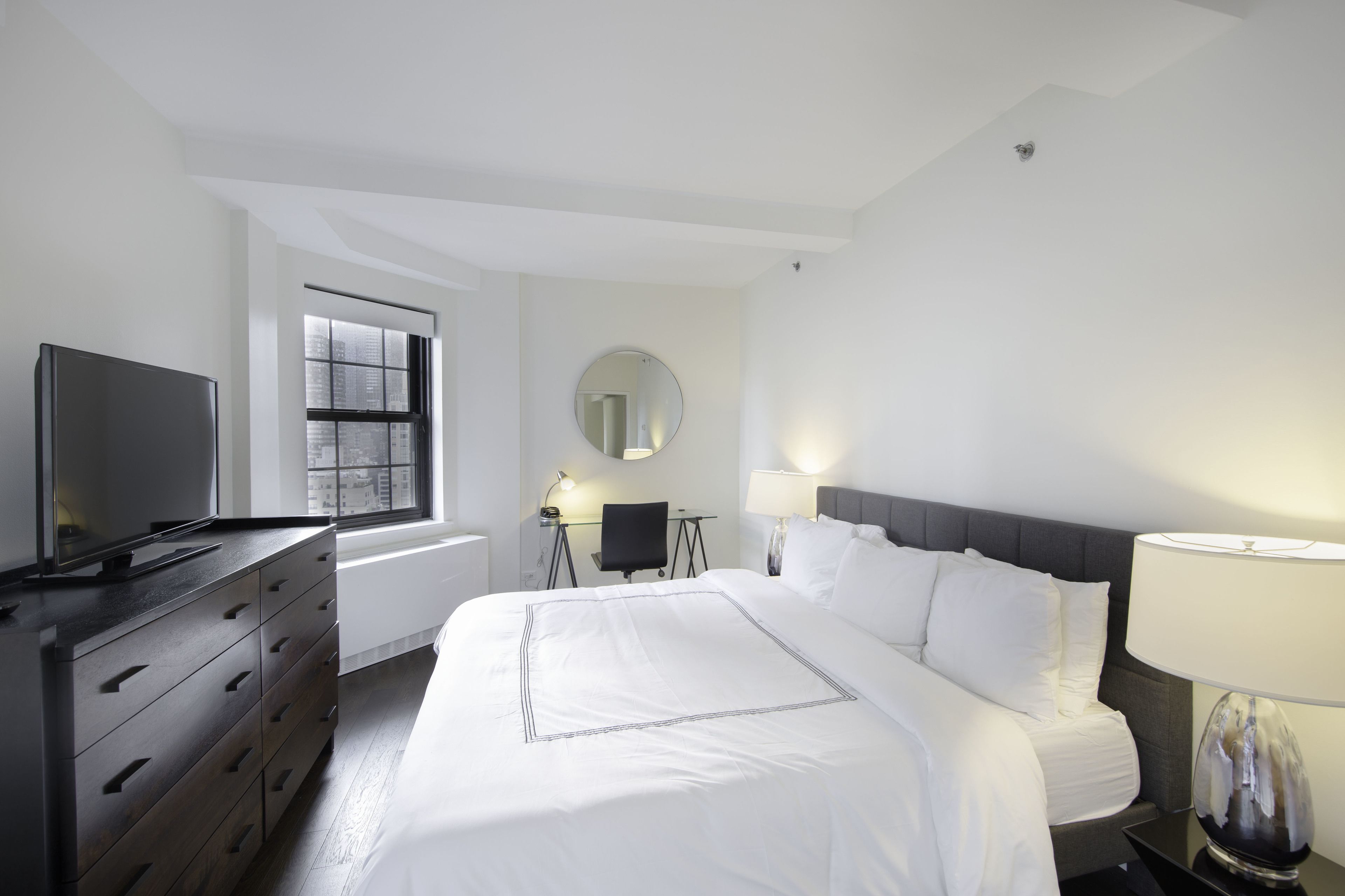 A hotel room with a bed, a TV, a desk with a chair, a round mirror, and a window with a view of buildings.