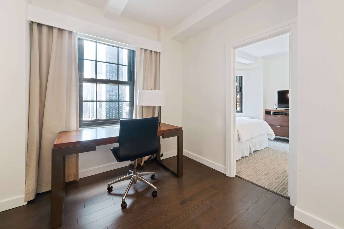 A hotel room with a wooden desk, a chair, a lamp, a bed with white bedding, and a view of the city through the window.