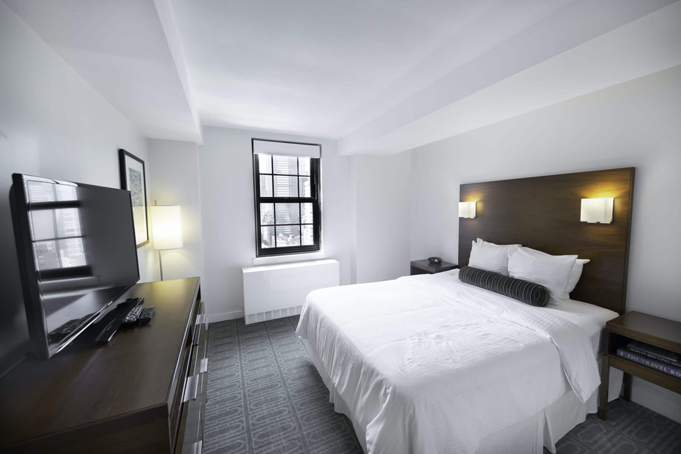 Habitación de hotel con una cama grande, mesitas de noche, un escritorio con computadora, un televisor y una ventana con persianas.