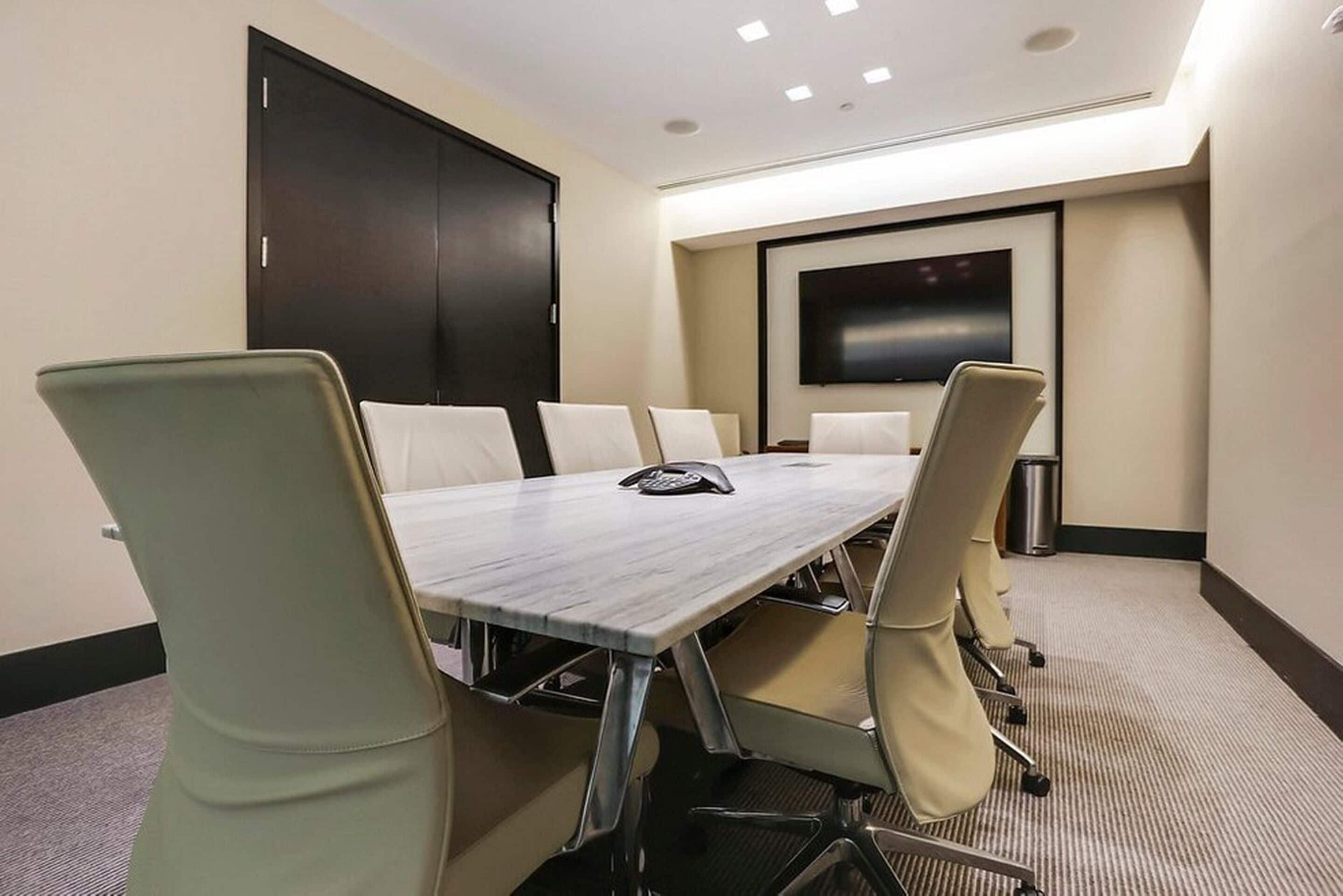 A conference room with a long table, six chairs, a projector screen, and a trash can.