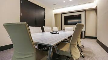 A conference room with a long table, six chairs, a projector screen, and a trash can.