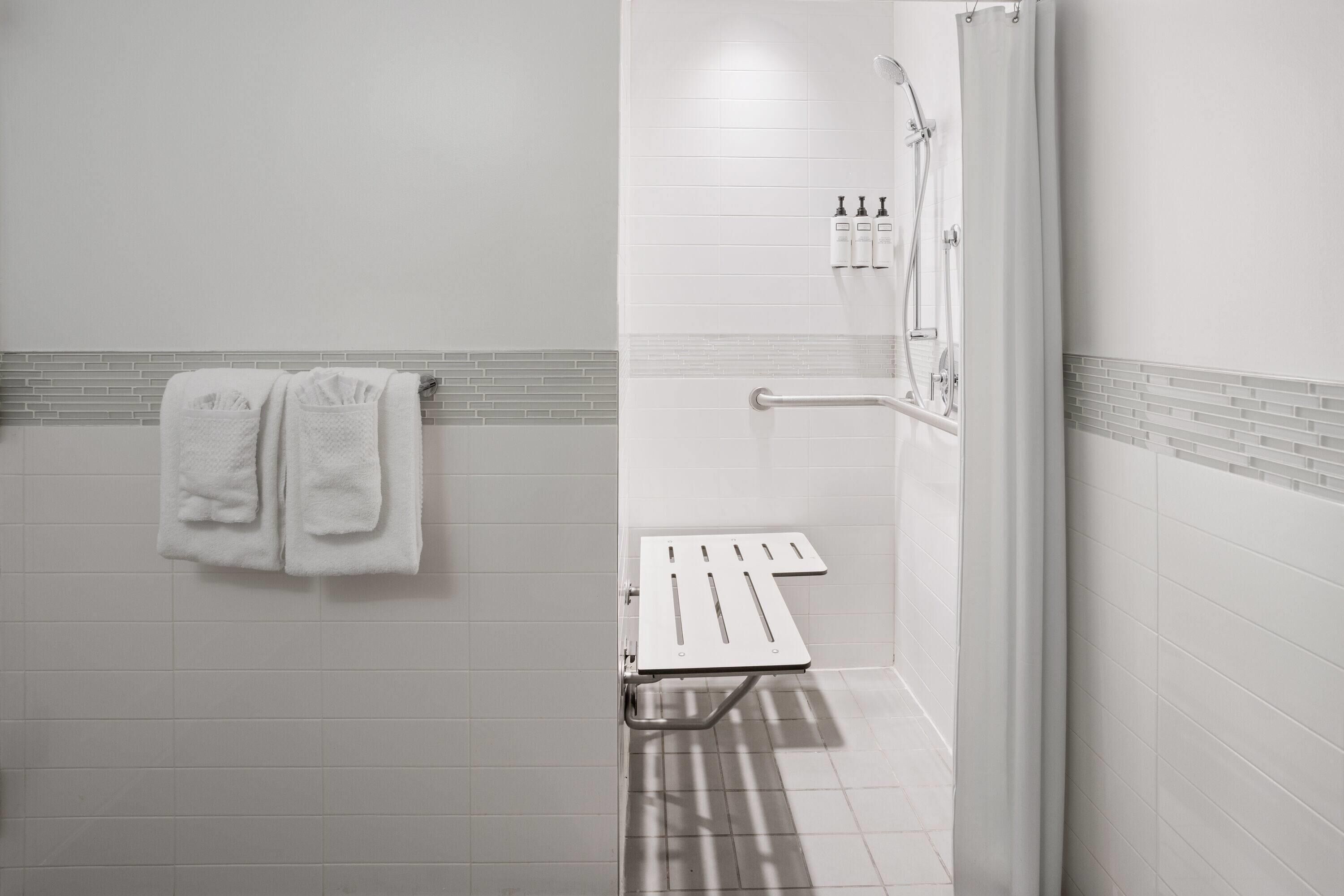 A bathroom with white tiles, a towel rack with two towels, a shower with a handheld showerhead, and a grab bar.