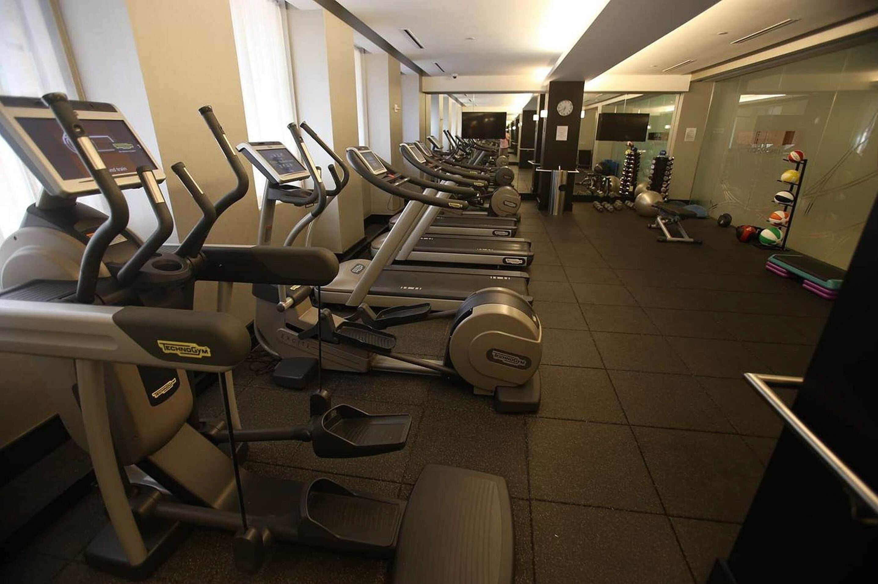 Fila de bicicletas estáticas y cintas de correr en un área de gimnasio, con pelotas de ejercicio y pesas a la derecha.